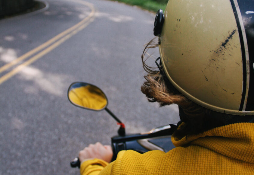 Osoba koja vozi motocikl ili skuter po zavojitoj cesti. Pogled je straga, obuhvaća gornji dio leđa vozača, kacigu i dio ceste ispred. Jakna je jarko žute boje.
